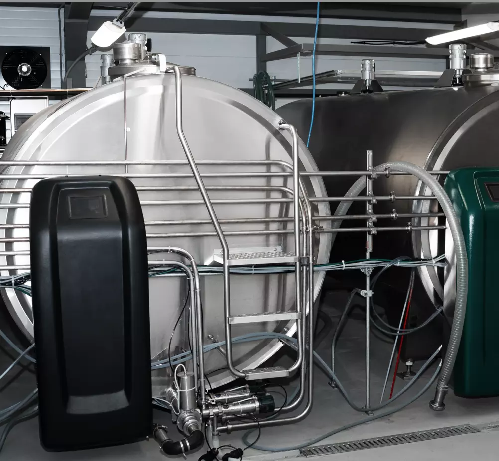 Industrial setting with two large stainless steel tanks connected by metal pipes and hoses. A black and green control panel is visible. The scene conveys efficiency and precision.