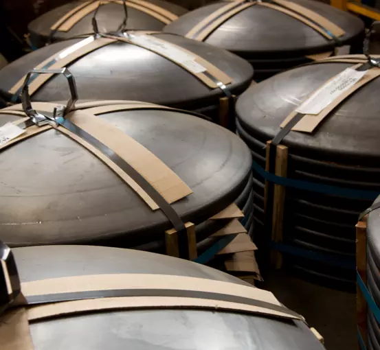 Stacks of rounded metal lids secured with blue straps and cardboard on top, arranged in an industrial setting.