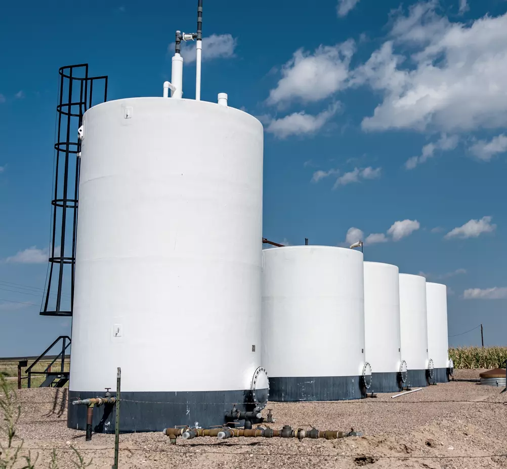 Row of large, white industrial storage tanks under a bright blue sky with scattered clouds. The tanks are set on gravel, conveying an industrial setting.
